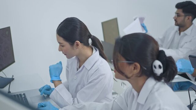 Asian woman in lab coat discussing work with colleague at desk with monitors, male researcher in background, friendly lab team conversation.