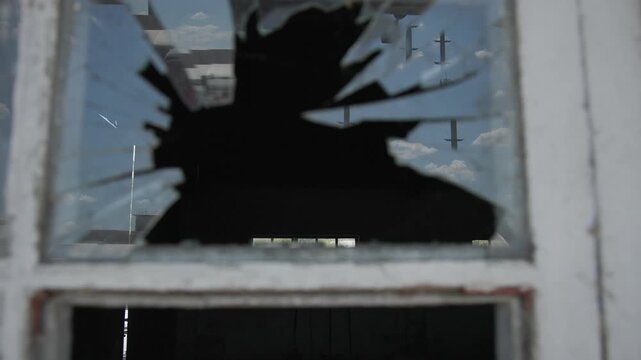 Close-up of a shattered window in an old abandoned building, revealing jagged glass fragments and weathered wooden frames. The damaged pane creates a dramatic urban decay scene, symbolizing neglect