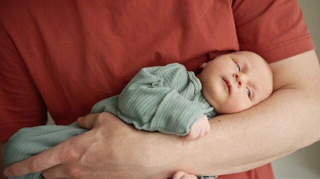 Young devoted father gently holding his newborn son in a warm embrace, creating a beautiful moment of family bonding. The infant looks around and then falls asleep in his dad's secure arms