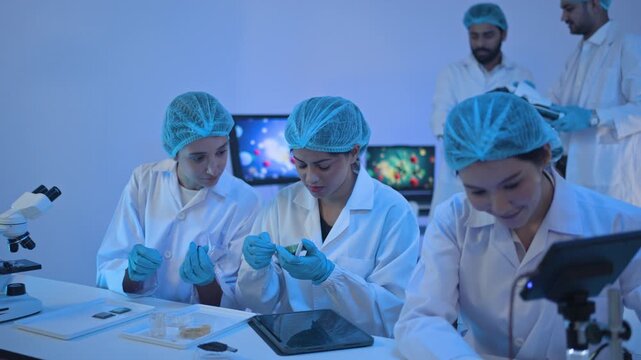Scientists in hairnets examining samples at bench with microscope and petri dish while colleagues talk behind, molecular images on screens.