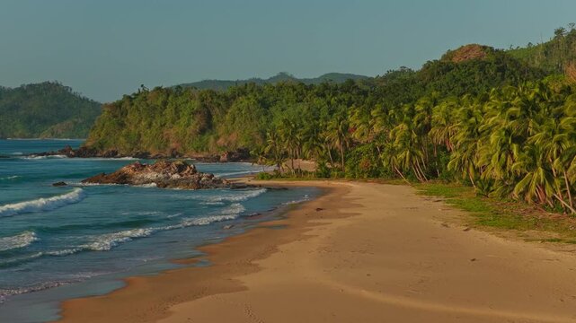 Aerial Backwards Reveal of Duli Beach in El Nido, Philippines. Telephoto Drone Pull Away View of Golden Sand Shoreline, Rolling Ocean Surf Waves, and Jungle Canopy.