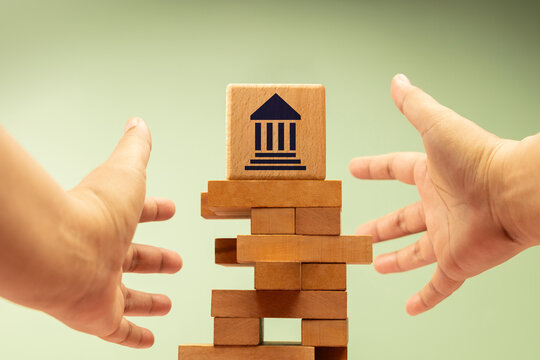 Hands reaching toward unstable wooden blocks with government bank symbol, economic finance uncertainty