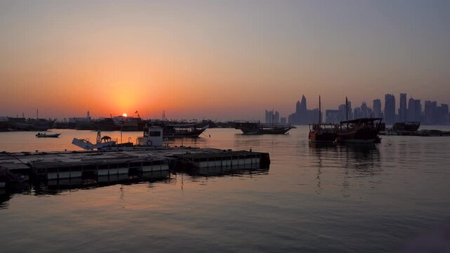 Stunning sunrise shot over Doha harbor featuring traditional dhow boats, a floating dock, and the hazy West Bay skyline silhouetted against a vivid orange and pink glowing horizon.
