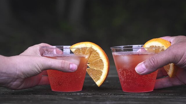 Slow motion two hands placing refreshing red cocktails with orange slices and ice cubes on a rustic wooden table. People toasting for a celebration or party