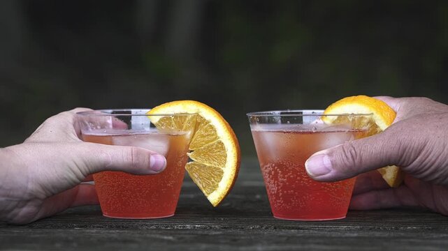 Two hands picking up plastic cups with orange-garnished cocktails from a wooden table. Friends making a celebratory toast outdoors