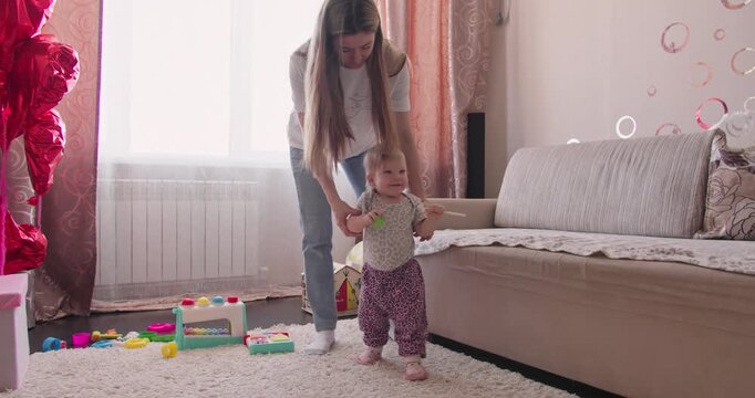 Mother supporting a toddler during early walking practice at home. First independent steps and child development in a family environment