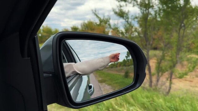 Woman hand waving in car back mirror riding countryside summer highway closeup. Female arm relaxing at road trip auto travel destination exploration discovery adventure. Expressway weekend vacation