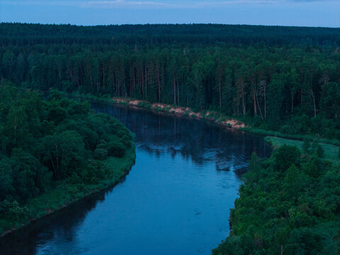 Aerial view of Gauja National Park featuring the winding Gauja River flowing through dense pine forests under a twilight sky in Incukalns, Sigulda Municipality, Latvia.