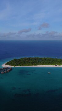 Aerial daylight pan shows a Maldivian coral atoll resort, crescent beach, palms, wooden jetty, overwater villas, reef patches, a drifting dhoni, and moving cloud shadows.