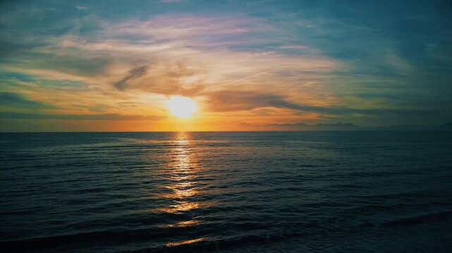 A breathtaking panoramic view of a dramatic golden sunset over the Mediterranean Sea in Agropoli, Cilento coast. The sky is filled with vibrant hues of orange, pink, yellow, and blue clouds, while the
