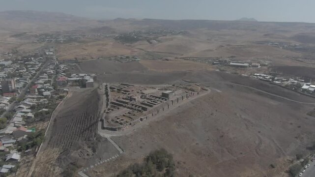 An aerial view beautifully showcases the ancient Erebuni Fortress, a historic Urartian fortified city situated on Arin Berd hill in Yerevan, Armenia, surrounded by the modern urban cityscape.

