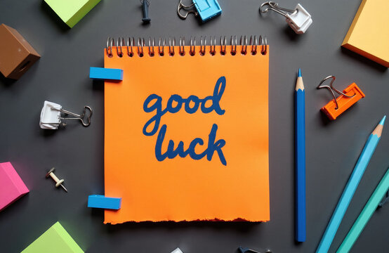 Handwritten good luck message on orange paper. Desk with stationery items, colored pencils, binder clips and colorful sticky notes. Office or school supplies background.