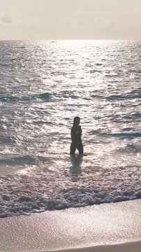 A woman walks from shallow surf on a Maldives resort beach, pauses hands behind head, waves lap at her feet, tiny boat and islet on horizon in golden hour light.