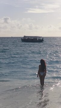 Vertical seaside scene in Maldives shows a woman in a white bikini wading toward a wooden dhoni as choppy waves glitter and the vessel gently rocks in backlight.