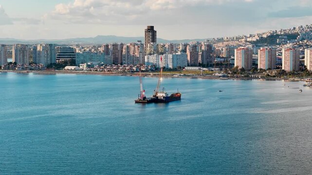 Floating crane barge and dredging vessel operating in bay water near dense city waterfront. Marine engineering work for seabed excavation and coastal water maintenance