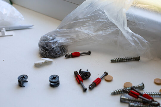 Various furniture hardware, metal screws, minifix cam dowels with red plastic parts, and screw caps are scattered on a white windowsill. The shot demonstrates fastening components and connectors used 