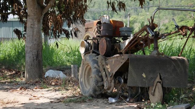 An old farm tractor stands in a grassy field beside a tree in the countryside. Rural heritage.