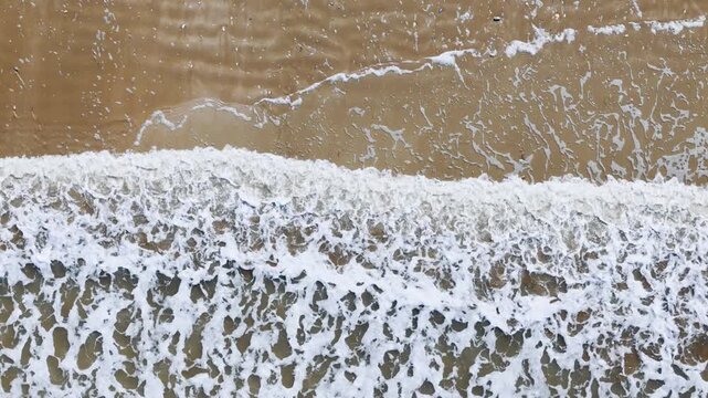 Breaking waves with white foam spreading across sandy beach, drone top down. Abstract shoreline pattern and sea texture