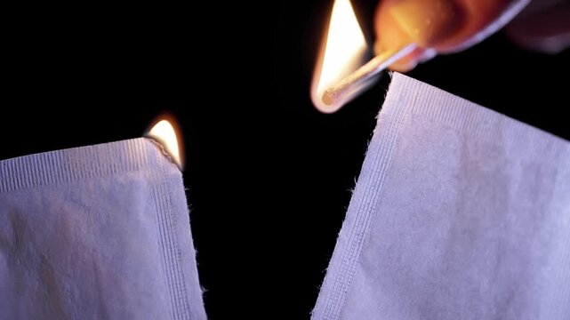 A hand holds a lit match to ignite the torn top edges of white paper packets in the dark. Concept of destruction, burning evidence, starting fires, and dangerous heat ignition. Slow motion.