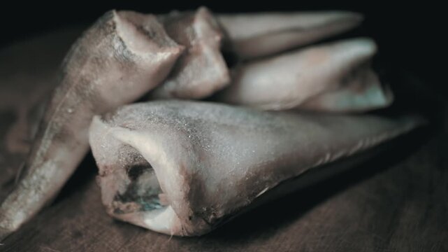 Frozen headless hake fish carcasses covered in ice crystals rest on a wooden cutting board. Concept of raw seafood preservation, cold storage, and healthy pescatarian meal preparation.