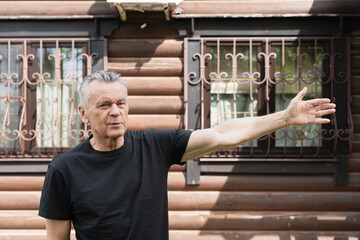 Fototapeta premium Senior man gesturing with one arm outdoors near a wooden house, concept of explaining, welcoming, guiding or presenting a place.