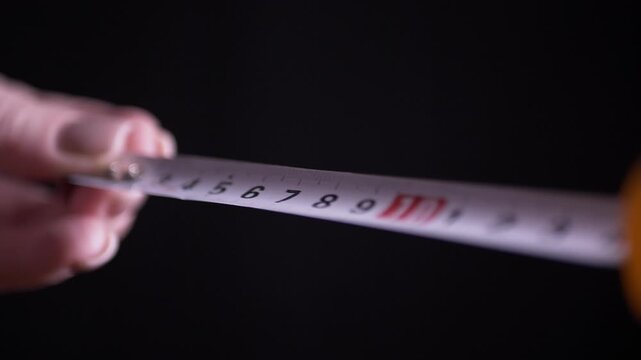 A macro view of hands slowly extending a yellow retractable measuring tape against a dark background. Concept of exact precision, careful planning, and professional home construction.