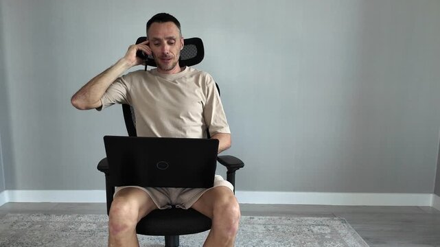 A young man suddenly appears in the frame, sitting in an ergonomic chair, simultaneously working on a laptop and talking on the phone. Concept: multitasking and busyness
