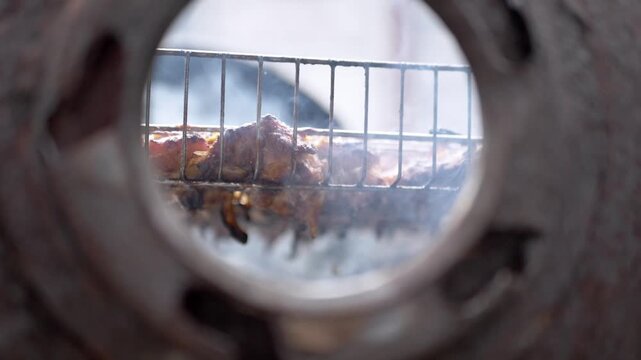 Succulent meat pieces roast on a metal grate inside a circular grill while thick smoke rises from the heat during the cooking process. Concept of traditional barbecue and authentic outdoor