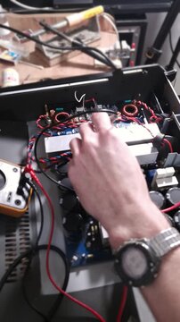 Close-up view shows a hand using a multimeter probe on an electronic circuit board. The repair process takes place on a cluttered workbench filled with various tools and components.