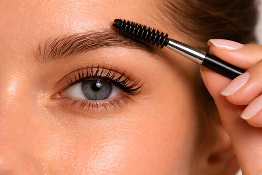 A close-up shot of a woman's eye and eyebrow as she uses a spoolie brush to groom and shape her brow hairs, highlighting natural beauty and makeup care.