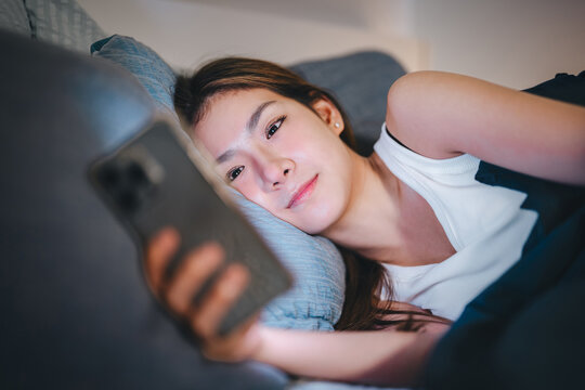 Young adult asian woman lying comfort on bed relaxing at night bedroom scrolling on phone for social media, digital dating online
