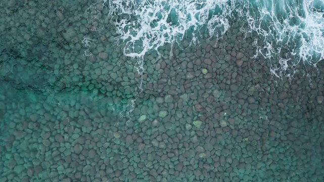 Aerial top view of ocean water moving over submerged stones, white foam forming natural patterns on rocky coastal seabed