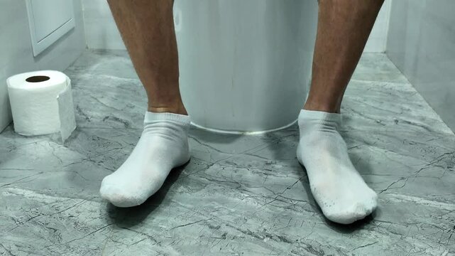 Close-up of a person's feet sitting on a toilet with a roll of toilet paper standing nearby