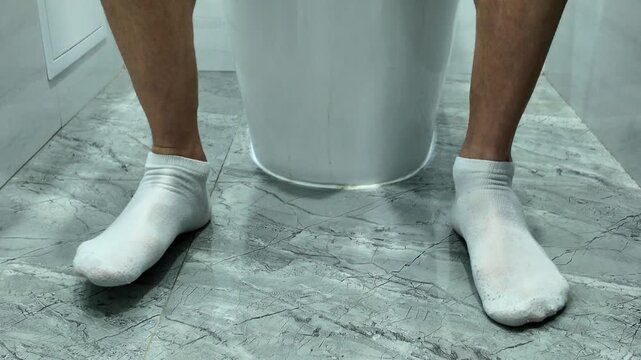 A man's feet in white socks tap the floor in boredom as he sits on the toilet. Concept of constipation or diarrhea