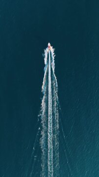 Vertical video. Aerial drone view of a fast motorboat in the blue ocean. Top down footage of a white boat cruising in turquoise sea water with waves and foam. Summer travel and vacation. 