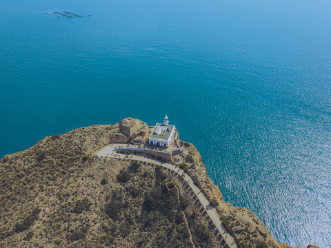 Aerial view of the Albir Lighthouse situated on a rugged cliff overlooking the Mediterranean Sea with a winding path and rocky terrain in L'Albir, Valencian Community, Spain.