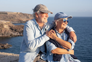 Fototapeta premium Senior couple embracing along the coastline at sunset, enjoying an active retirement vacation together while smiling and looking happily at the ocean view