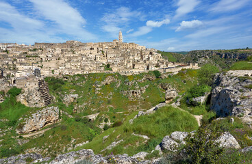 Fototapeta premium Panoramic view of the medieval cave city with historic stone architecture, dramatic cliffs and lush valley beneath a clear blue sky. Matera, Basilicata, Italy