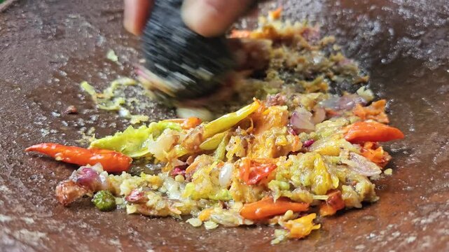 Close up of a person grinding red and green chilies onions and garlic using a traditional stone mortar and pestle to make a spicy chili paste