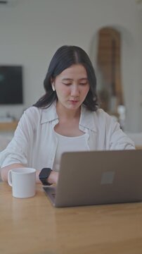 Young Asian woman using laptop with wireless earbuds for online meeting, working remotely from home at wooden table with coffee mug, talking with her team