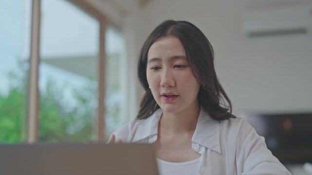 Asian woman using laptop for online video conference with her team, working remotely from home, talking and discussing work in a living room