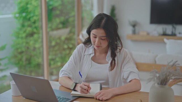 Asian woman taking notes by hand and planning her finances at home, writing in notebook beside laptop, organizing tasks and setting goals in a bright minimal room