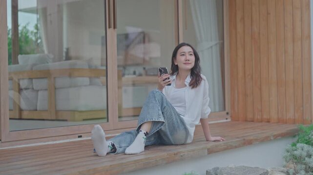 Asian woman sitting on wooden terrace holding smartphone, taking a break and relaxing at modern minimal home, enjoying slow living and a calm weekend moment