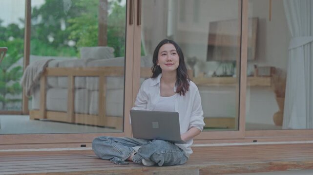 Asian woman sitting cross legged on wooden terrace using laptop, working from home in a calm minimal space, balancing work and relaxation in a slow living lifestyle
