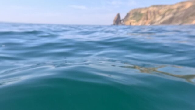 Ocean surface waves rippling blue water at sea level with blurry rocky cliffs in the distant background during a calm summer day