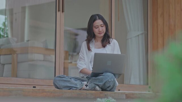 Asian woman sitting cross legged on wooden terrace working on laptop at modern minimal home, enjoying flexible remote work and a calm slow living lifestyle outdoors