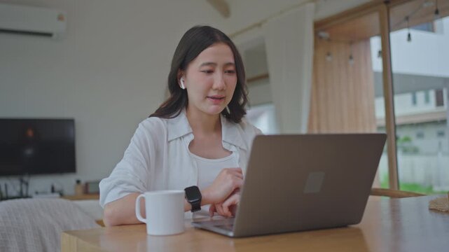 Asian woman wearing earbuds on online video call, working from home on laptop with AI assistant support, talking and typing during a remote meeting in a modern room