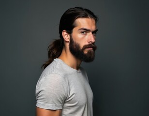 Fototapeta premium Man with long dark hair tied in a ponytail and full beard wears light grey t-shirt. He looks sideways with serious expression. Studio shot against dark grey background.