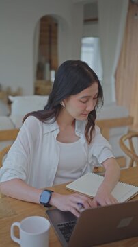 Asian woman wearing earbuds typing on laptop and writing notes in notebook, working from home and planning tasks, organizing ideas in a bright modern room