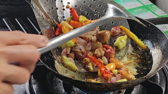 Person using a slotted spoon to scoop fried spicy chili peppers garlic and shallots from a hot frying pan